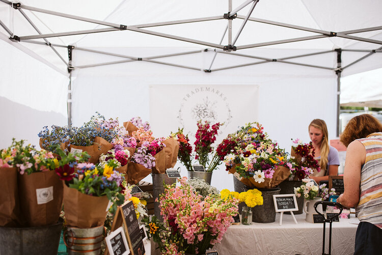 Wanting a profitable flower farm doesn't make you selfish. Jenny Marks is selling flowers at her local farmers market, her stand features the Trademarks Flowers logo in a white tent. The table is full of colorful flowers. A customer is purchasing.