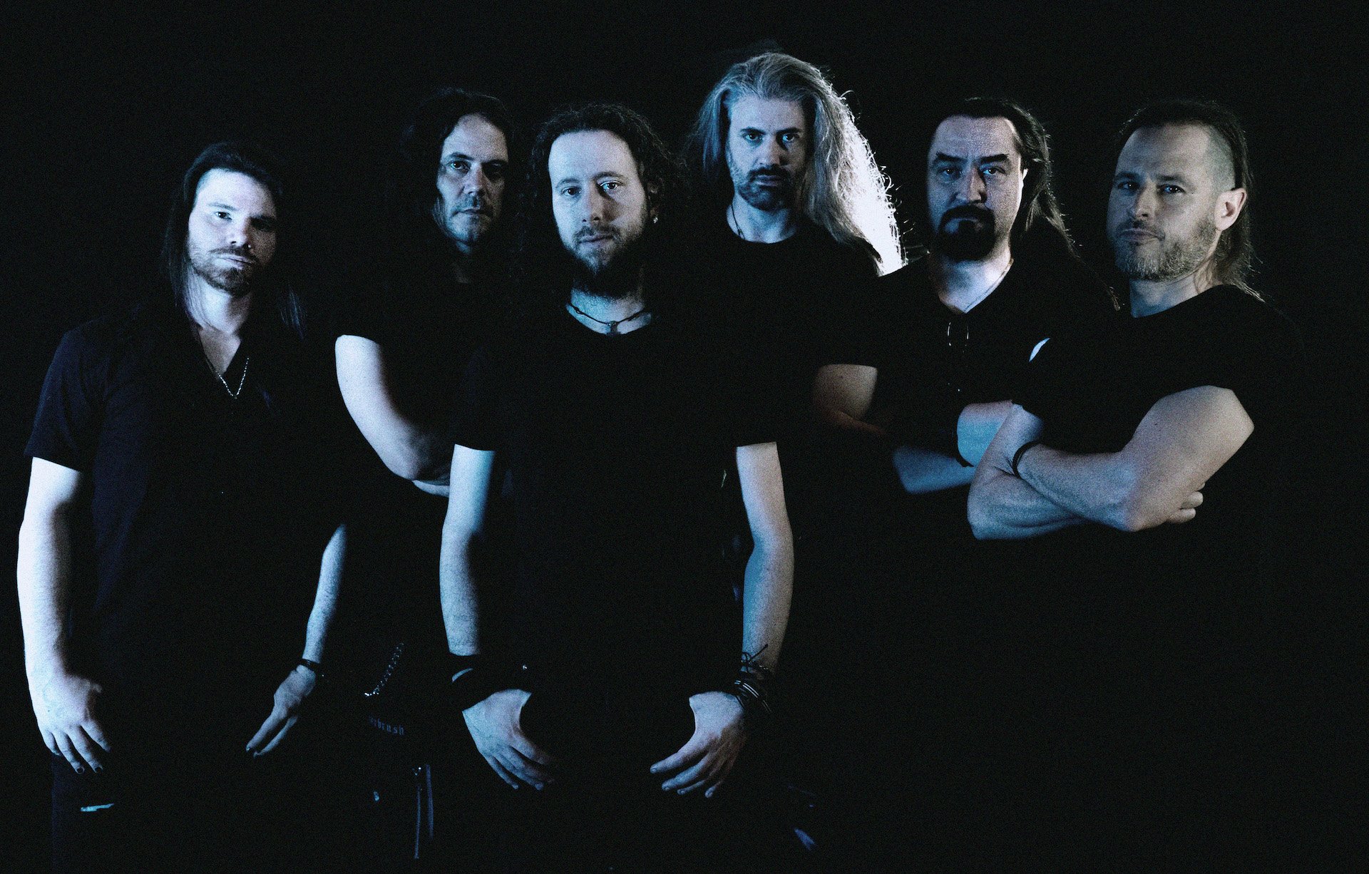 Group of six men in black clothing, standing against a dark background, with serious expressions.