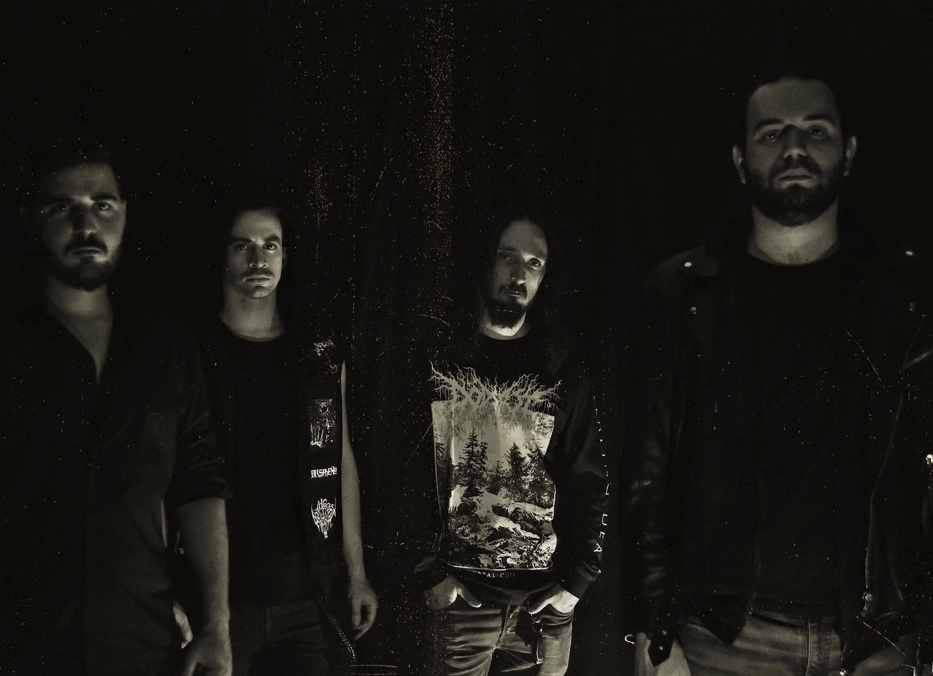 Four members of a band in a dark, grungy setting wearing casual and leather clothing.