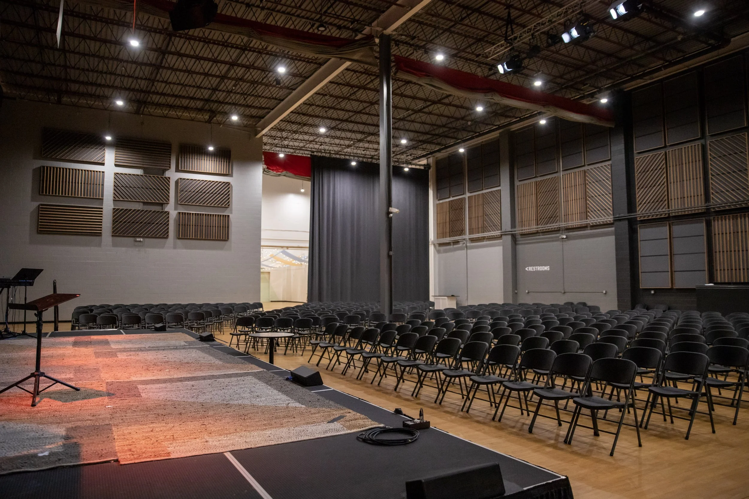 An auditorium space with rows of black chairs facing a stage with a microphone stand and a red and beige carpet in Richmond, VA. The ceiling is high with mounted lights and acoustic panels on the walls.