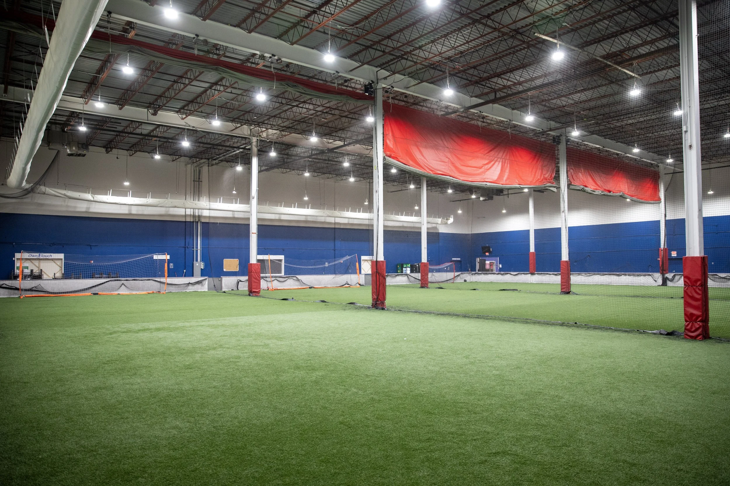 Indoor sports facility with artificial turf field, volleyball nets, and red banners hanging from the ceiling. Bright overhead lights illuminate the space.