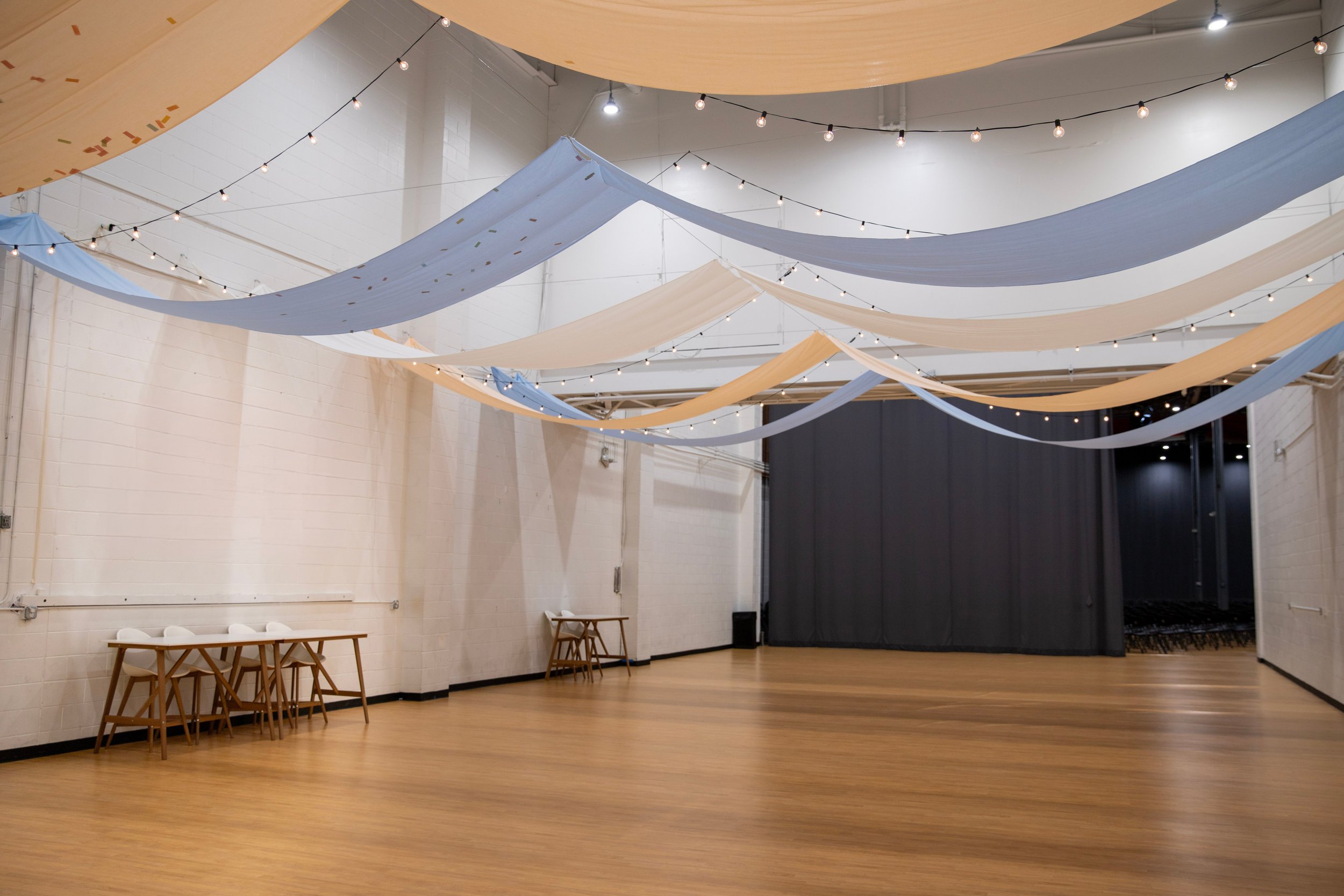 An indoor event space with draped fabric banners in light blue, cream, and beige hanging from the ceiling, string lights, a high ceiling, white brick walls, and a dark stage area in Richmond, Virginia.
