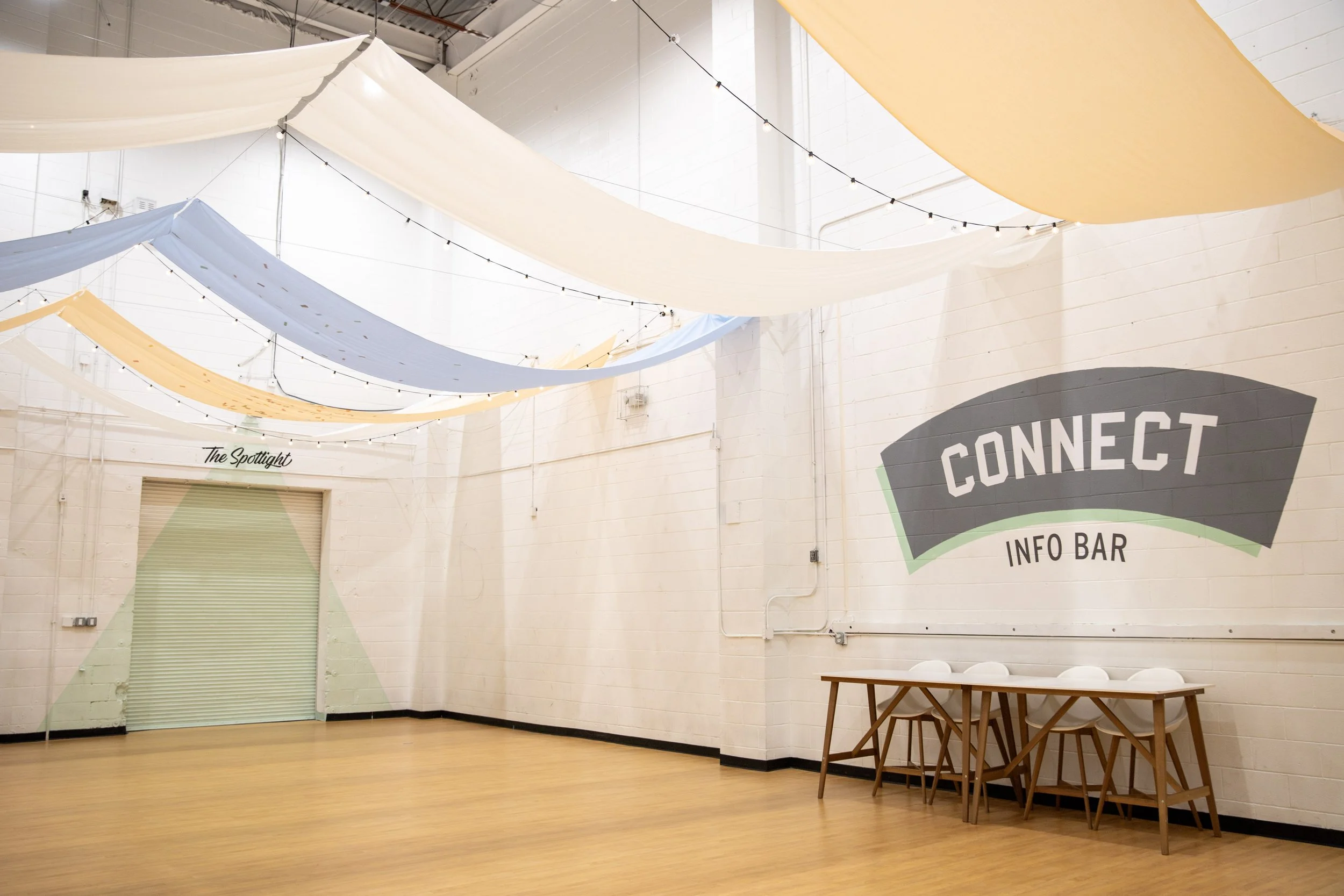 Interior of a large room with hanging fabric decorations in shades of cream, light blue, and yellow, string lights, a closed green roll-up door, a corner with a wall-mounted sign that reads 'The Spotlight,' and a table with chairs against a white brick wall with a 'Connect' sign.