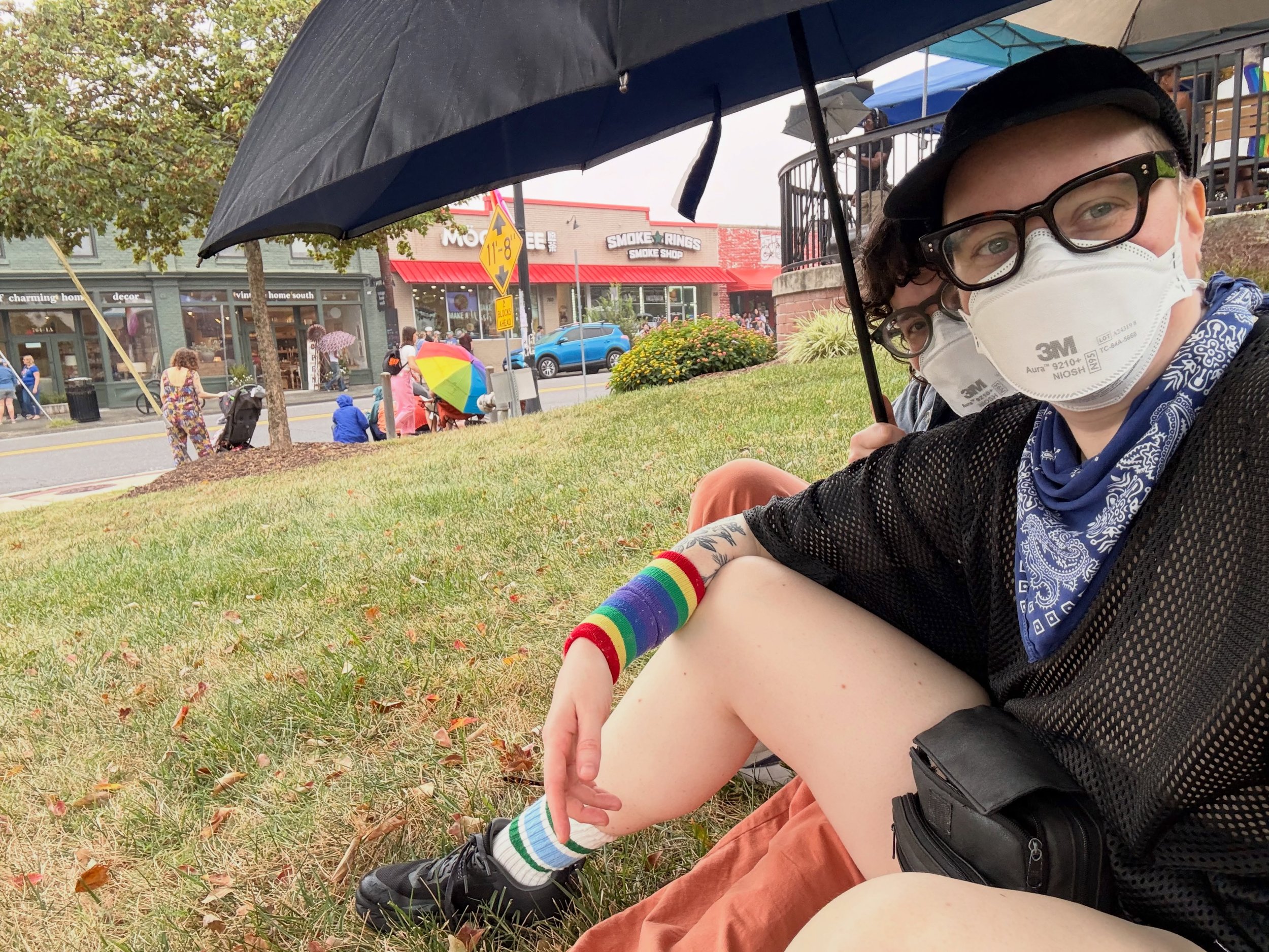 Rainy pride but we showed up! Thank you @lgbtqdurham for another hit parade! 🏳️&zwj;⚧️🏳️&zwj;🌈💘

Image description: two white people wearing n95 masks sit under an umbrella on grass with a street and parade watchers in the background waiting for 