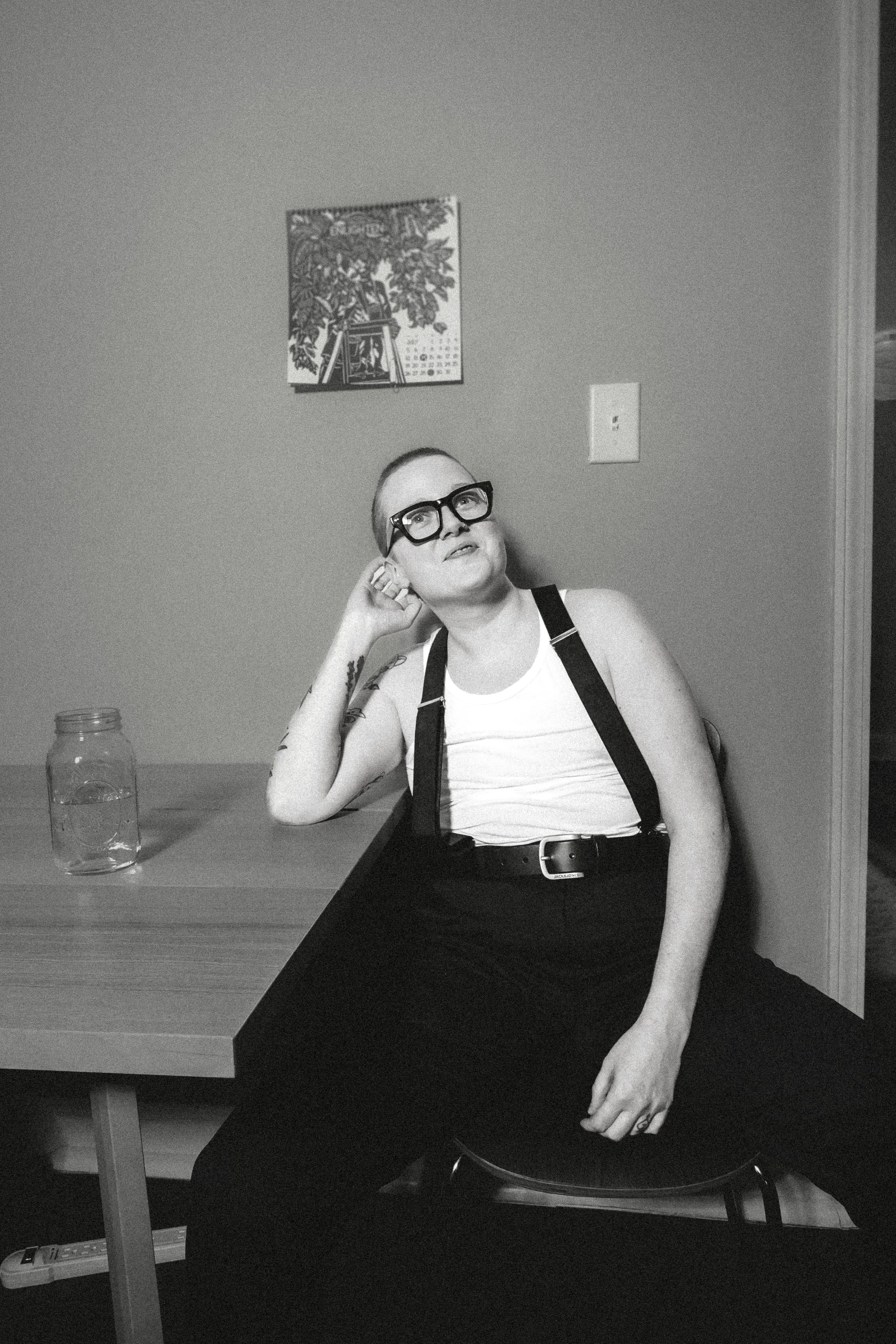 black and white photo of a white person sitting in a white tanktop with black suspenders and dark black glasses leaning their head on hand and looking up dreamily