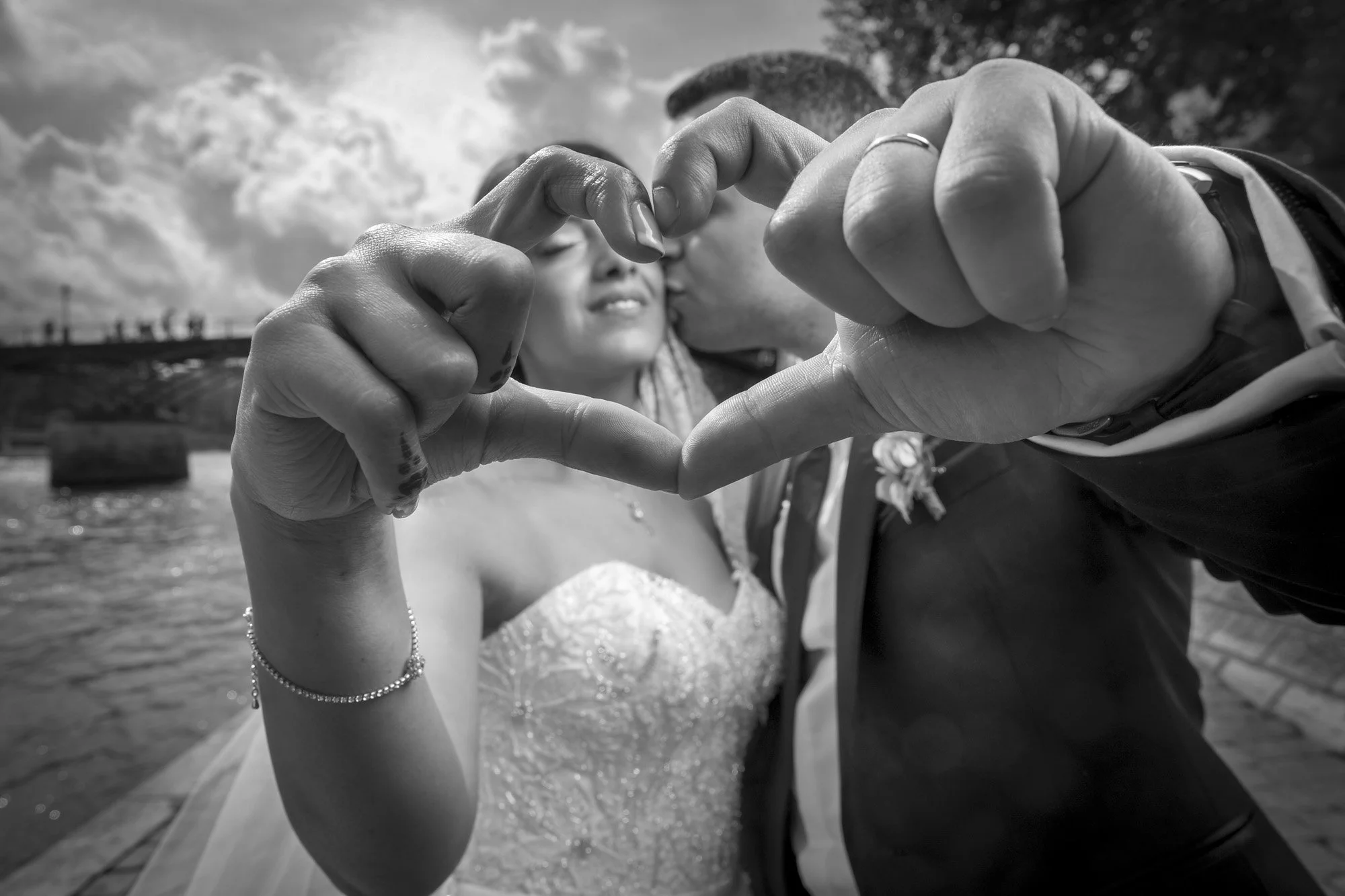 Un couple de mariés forme un cœur avec leurs mains devant l'objectif, avec un fond de ciel nuageux et les quais de Seine à Paris