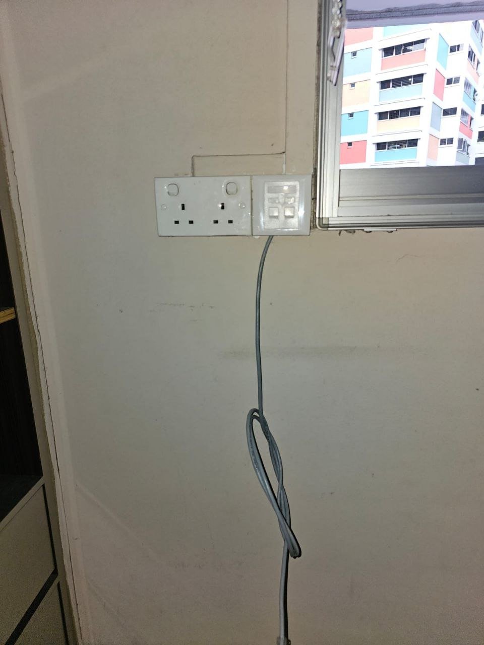 Power outlet with two sockets, a switch panel with four buttons, and a loose gray electrical cord hanging from the outlet, located near a window with a view of a colorful building outside.