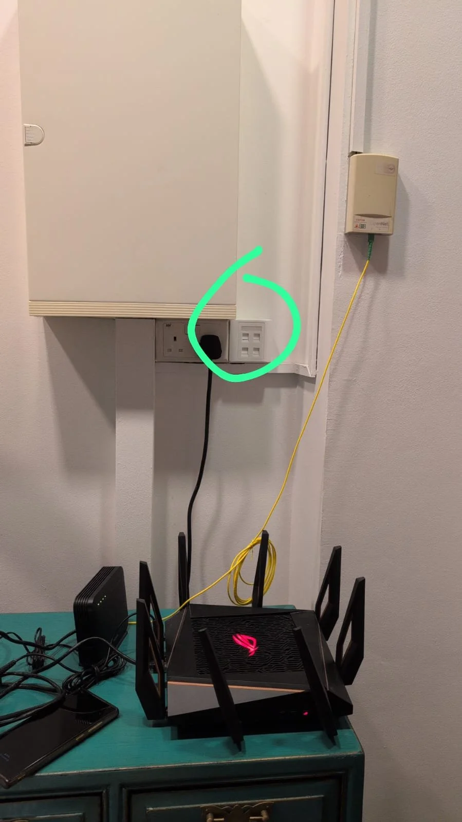 A router with multiple antennas on a table, with power and internet cables connected. Behind, a wall with an electrical outlet, a power cord plugged in, and a wall-mounted device with a yellow ethernet cable connected.