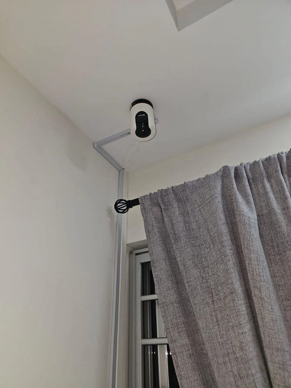 A ceiling-mounted security camera installed in a room near the corner, with a gray curtain hanging beside a window.