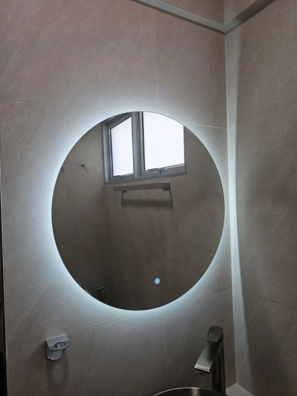 A round mirror with built-in lighting on a bathroom wall reflecting a window with frosted glass and a towel rack.