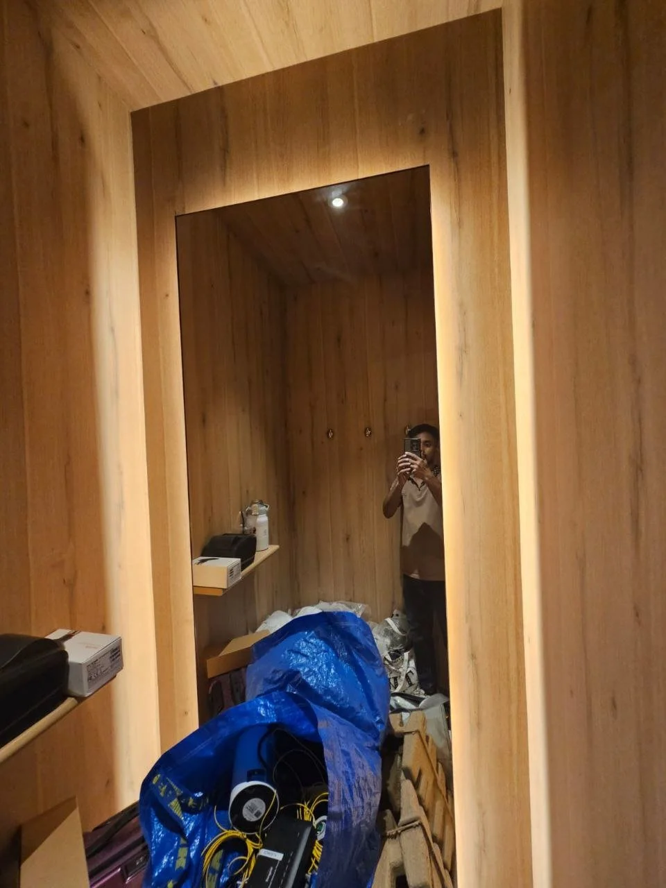 A cluttered room with wooden walls, a large mirror, and various items including a blue bag, headphones, and boxes. A person is taking a photo with their phone, reflected in the mirror.