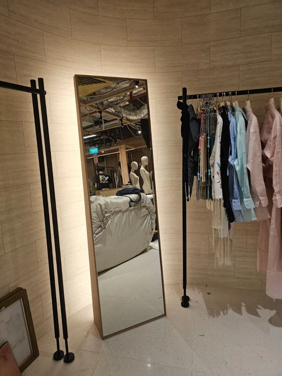 A full-length mirror leaning against a wall inside a store, with a clothing rack containing various shirts and necklaces nearby. The reflection shows mannequins and store ceiling.