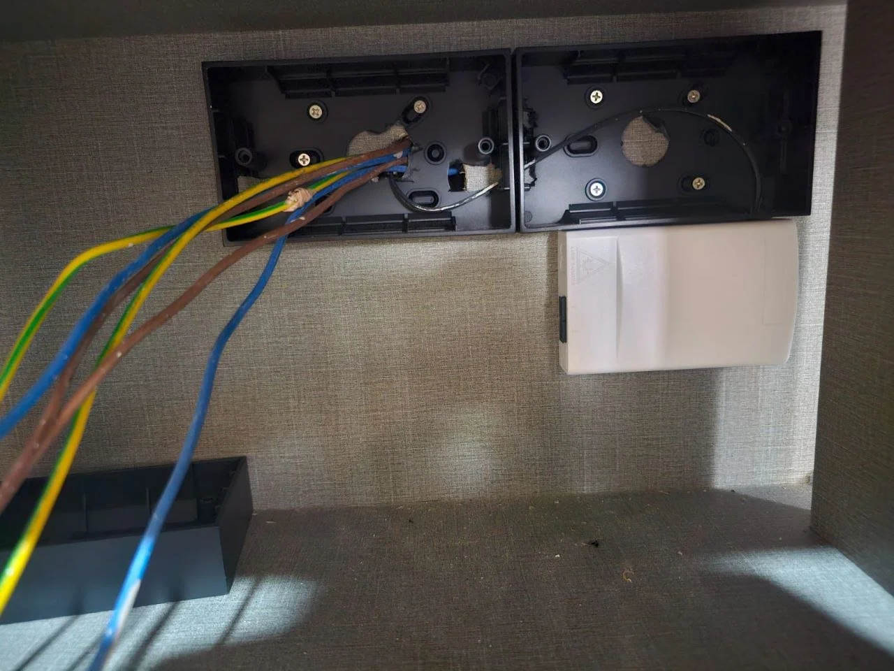Inside a disassembled electrical device box with several colorful wires and a white unused cover plate mounted on a textured wall.
