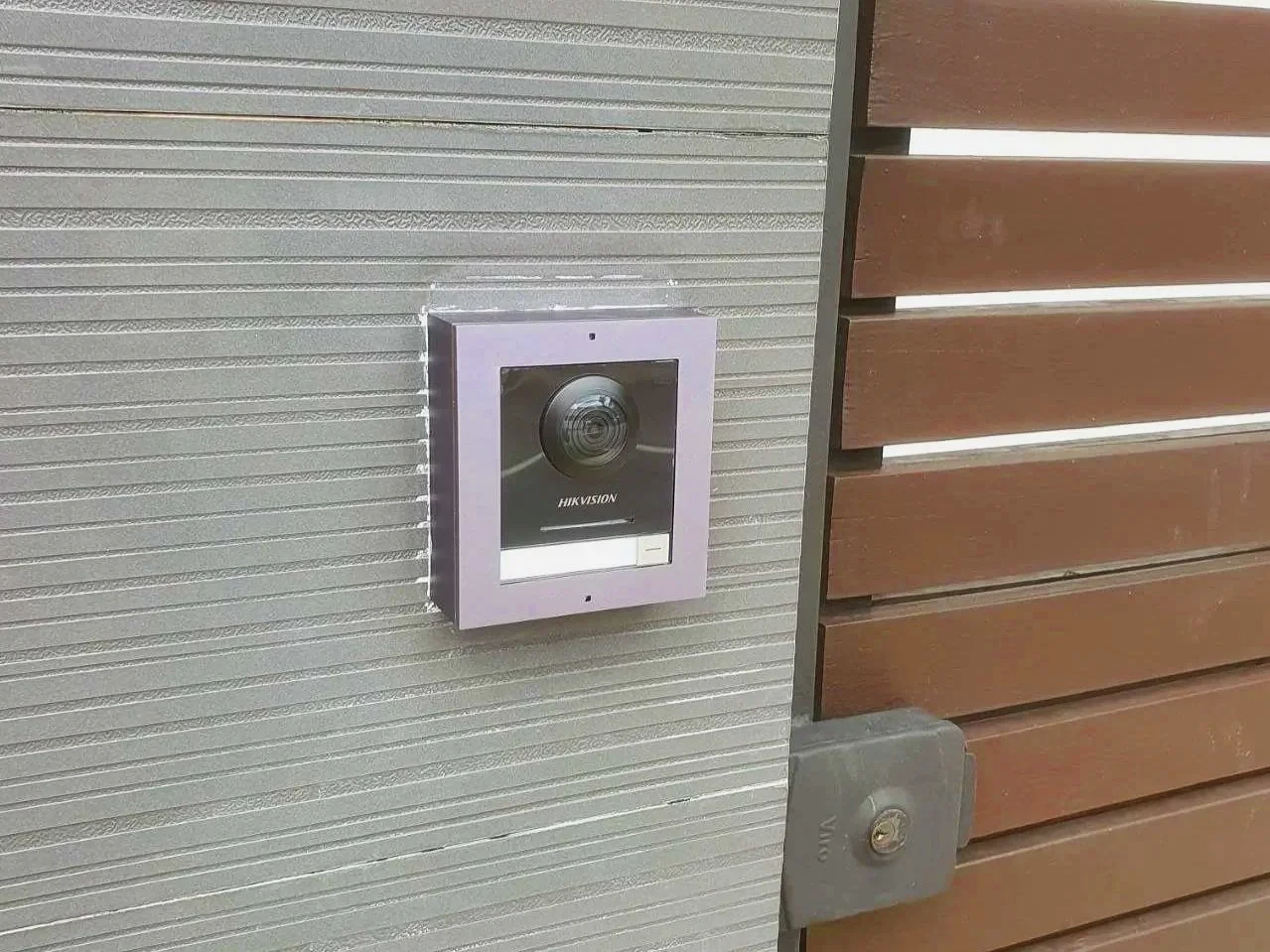 A doorbell camera mounted on a gray textured wall next to a wooden door with an electrical box below it.