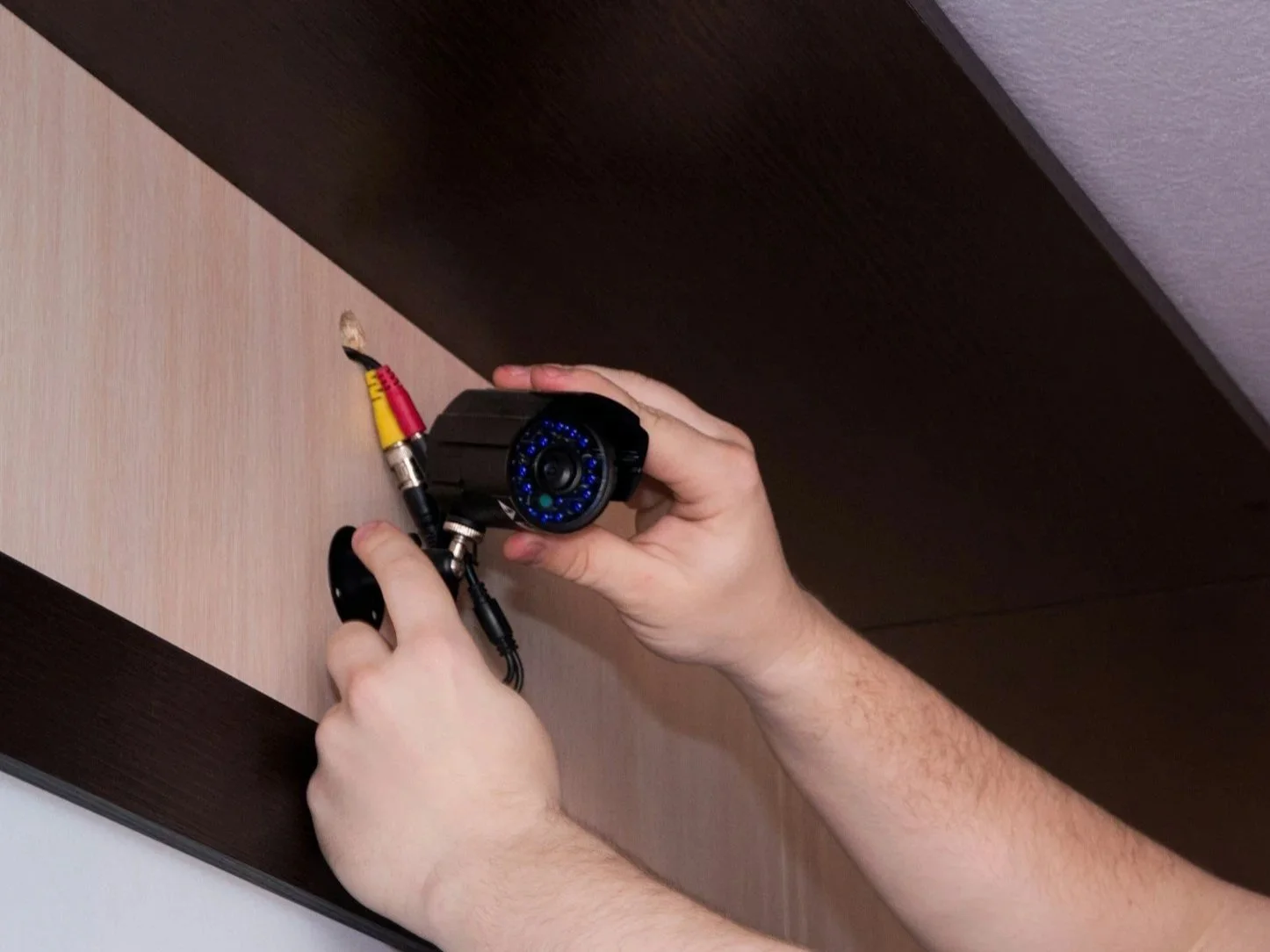 A person installing a security camera with a black body and blue LED lights onto a wooden surface using a screwdriver. The camera has a yellow, red, and black cable connected to it.