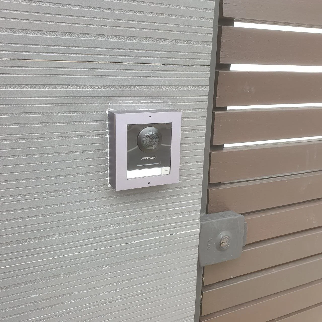 Close-up of a video doorbell mounted on a textured wall beside a brown wooden slat fence.