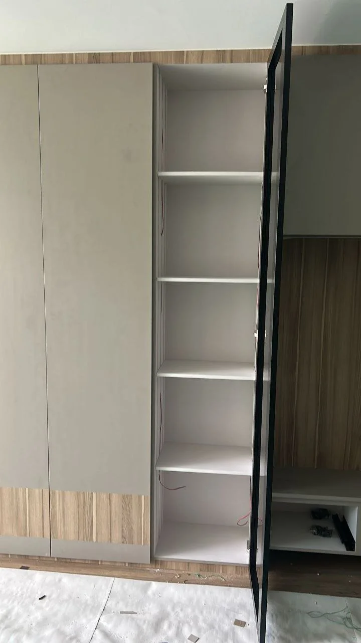Empty vertical cabinet with five white shelves and a glass door, next to a closed beige cabinet and a dark wood-paneled wall.