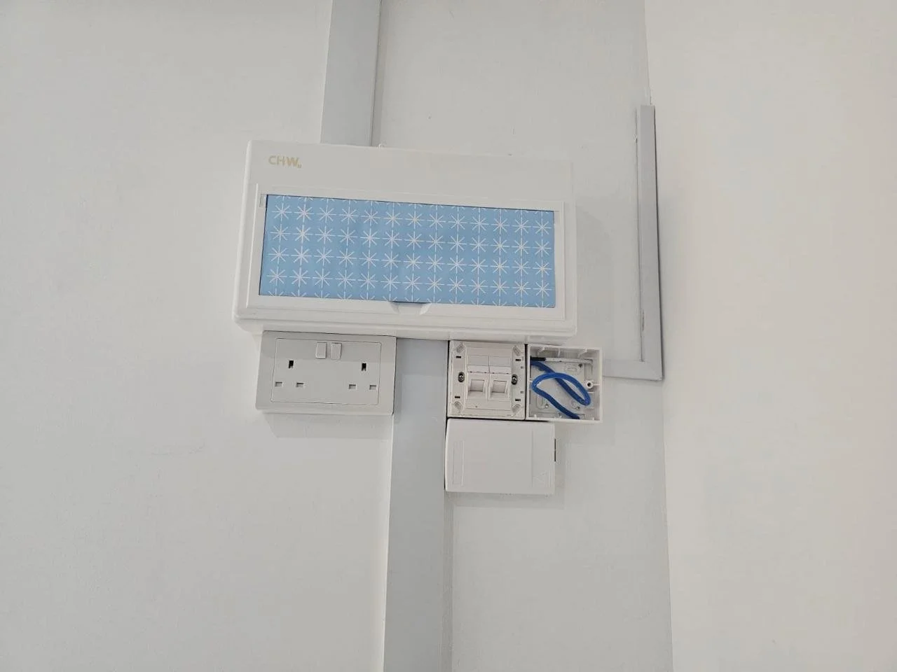 Electrical panel with a blue and white patterned cover, outlet, circuit breakers, blue wire cutters, and a white switch, mounted on a white wall.