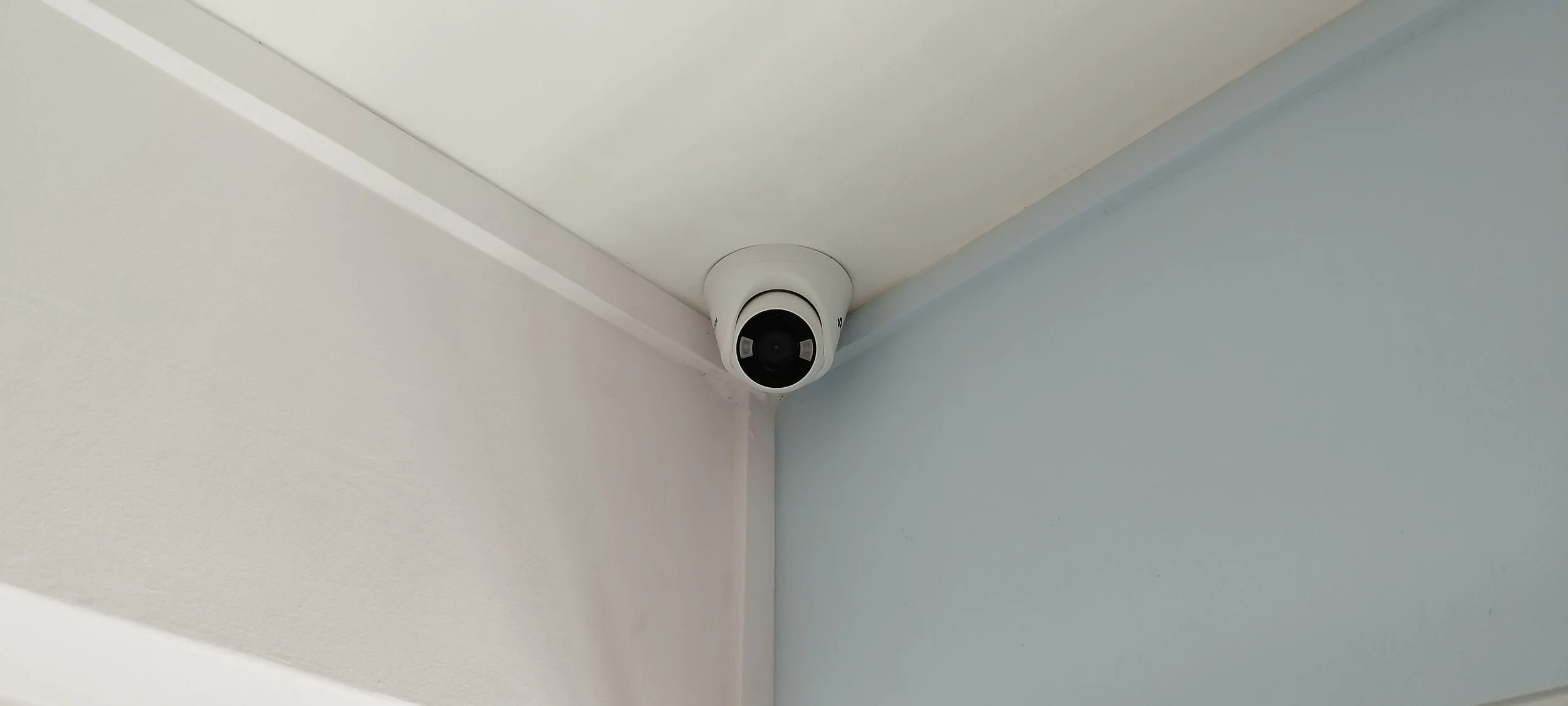 A white security camera mounted on the corner of the ceiling where two walls meet, one wall painted white and the other light blue.