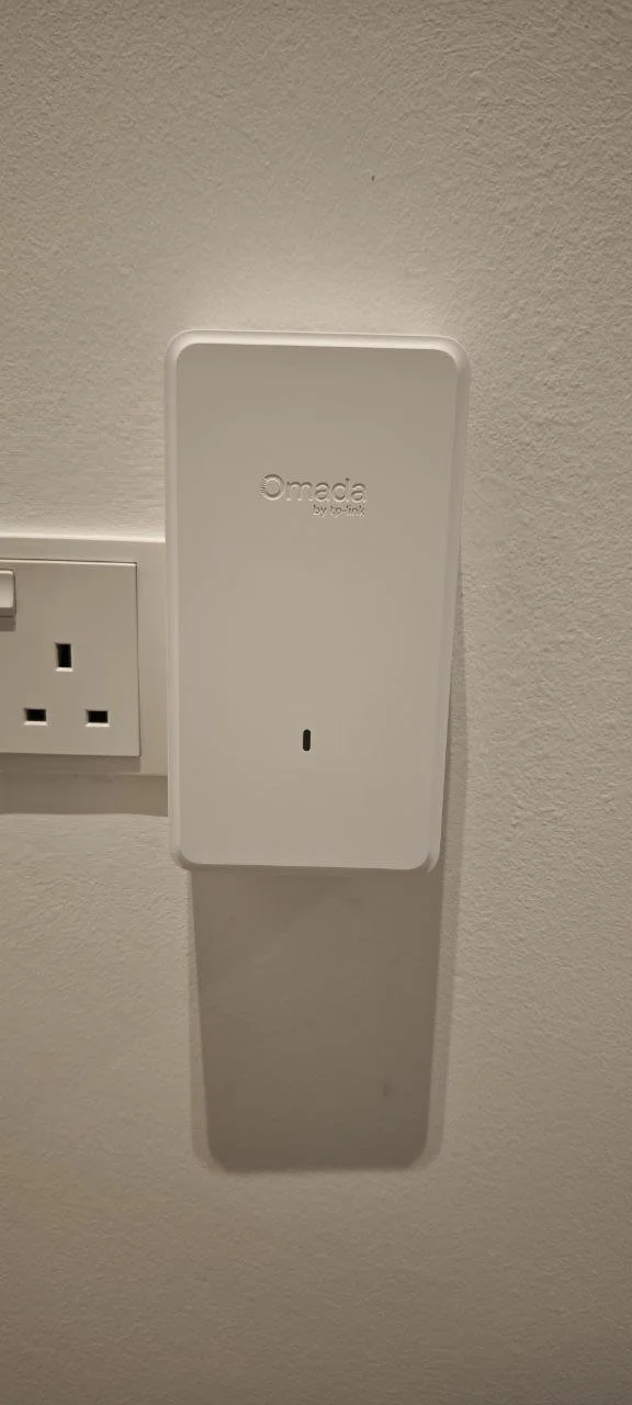 A white Omada Wi-Fi access point mounted on a wall beside an electrical outlet.