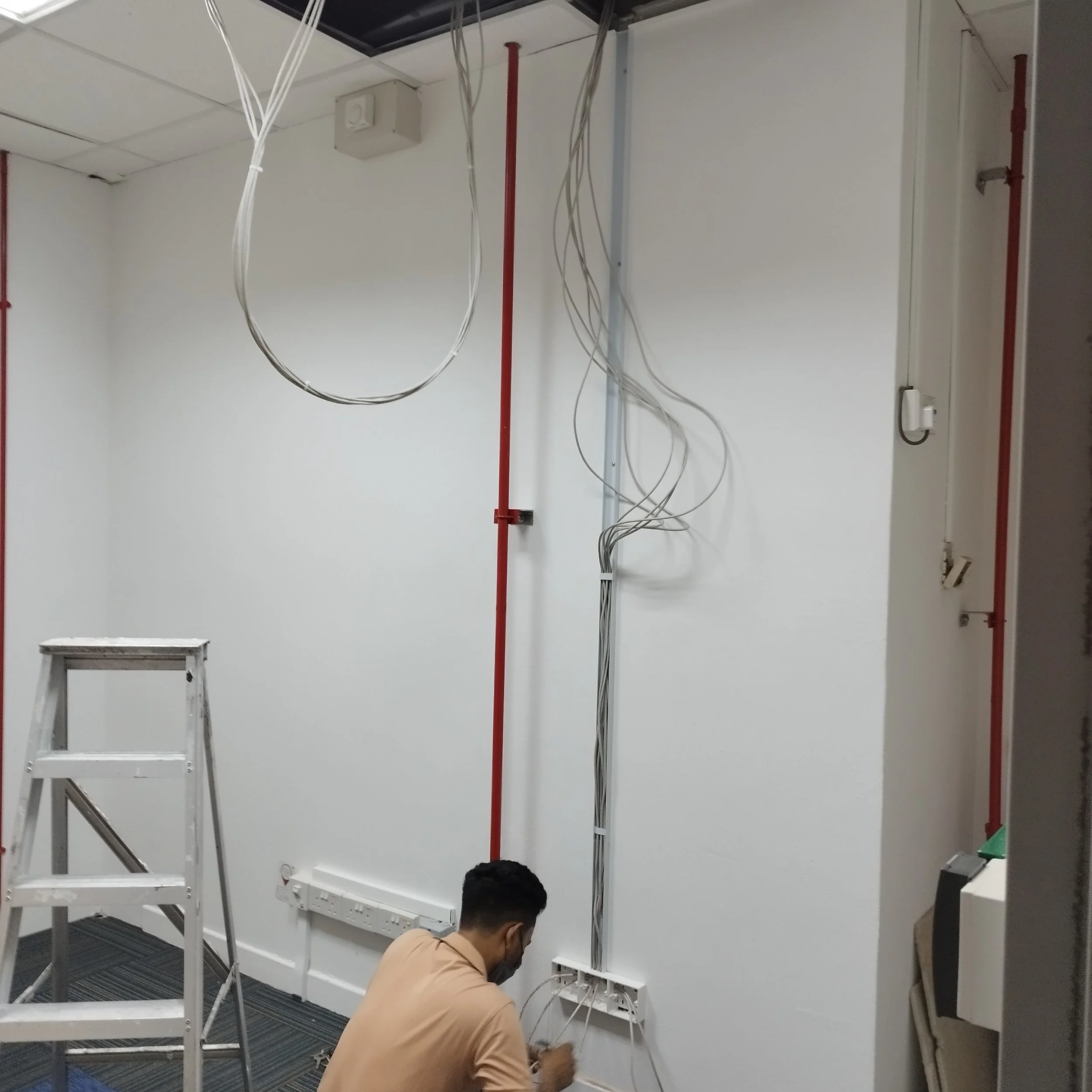 Two men working on installing electrical wiring in an office or server room with white walls, a ladder, and red conduit pipes.