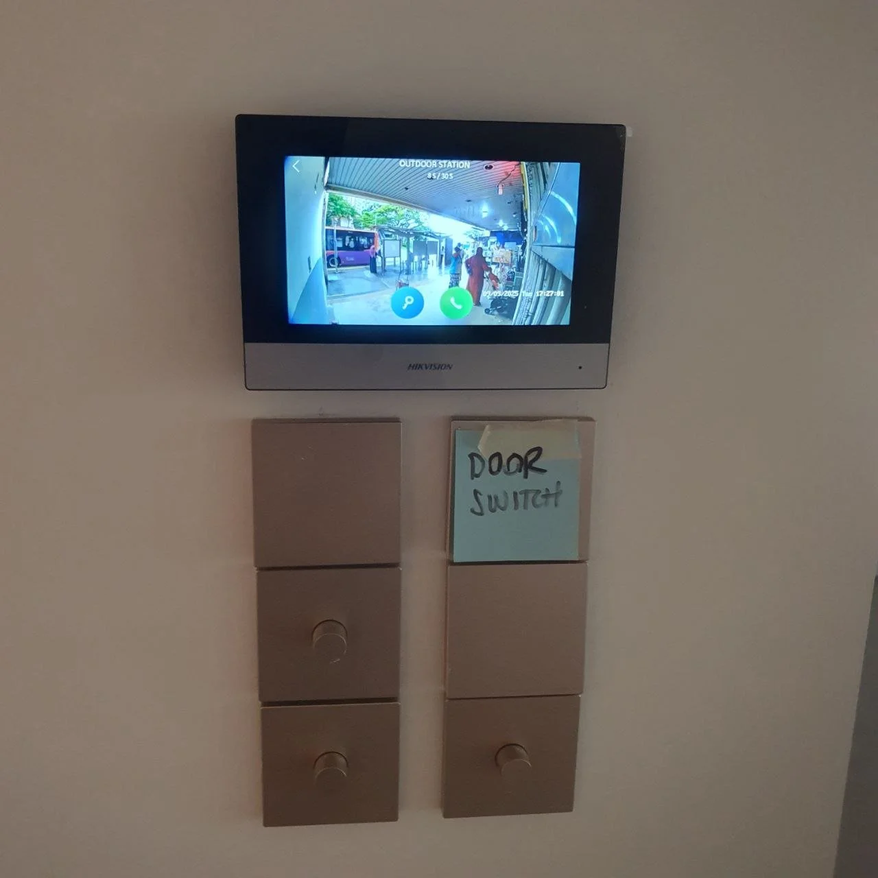 Wall-mounted video intercom screen showing a view of a bus stop with people walking and waiting, with a note labeled "DOOR SWITCH" written on blue paper below the screen.