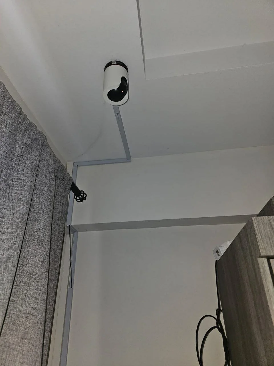 Close-up of a ceiling corner with a security camera installed and electrical wiring visible, next to a gray curtain and part of a wooden desk.
