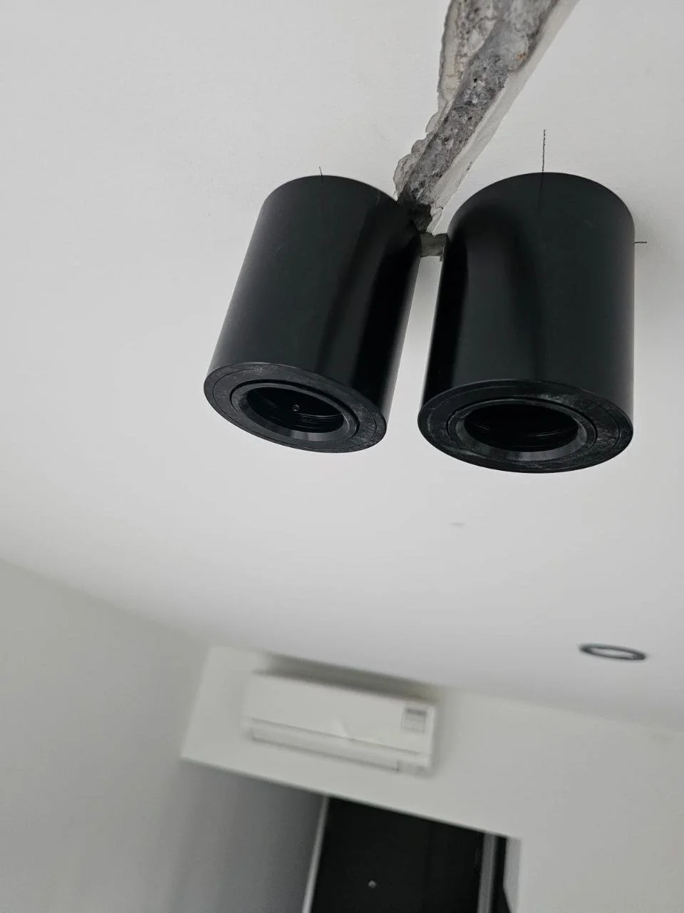Two black ceiling light fixtures with round cylindrical shapes mounted on a white ceiling, with a visible exposed beam and a wall-mounted air conditioning unit in the background.