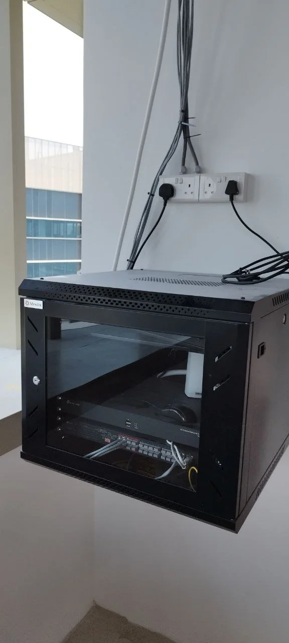A black metal server rack or cabinet with a glass door, placed on a white table in an office or server room. There are electrical outlets and cables on the wall behind it, some of which are connected to the server cabinet.