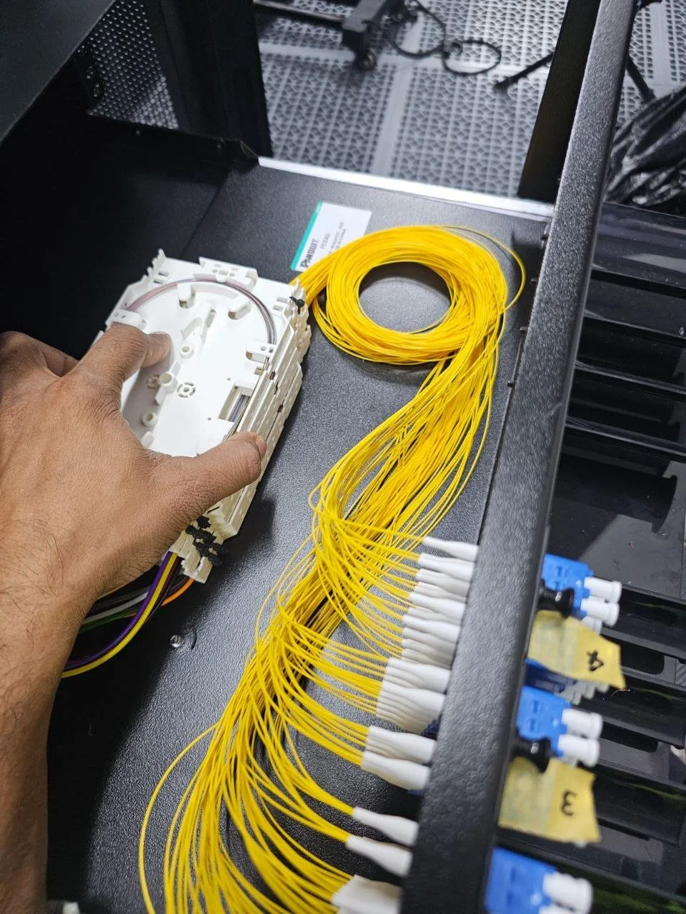 Hand installing or inspecting fiber optic splicing tray and yellow fiber optic cables inside a network cabinet or server rack.