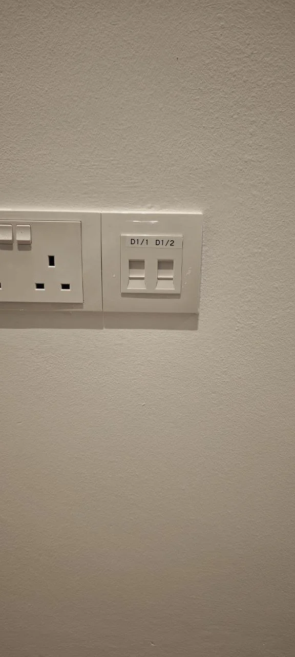 Close-up of electrical outlets and switches on a white wall, including a power socket and a switch labeled D1/1 and D1/2.