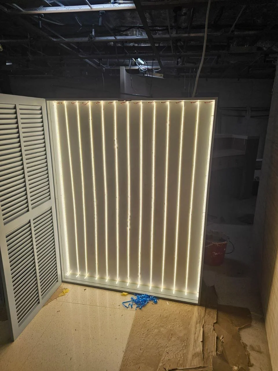 A partially constructed room with a neon-lit wooden slatted wall, an adjacent gray louvered door, and exposed ceiling wiring. Construction materials are visible on the floor.