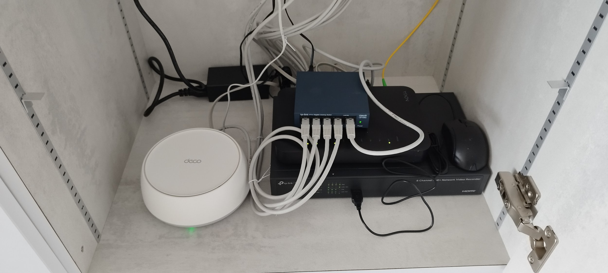 Electrical and networking equipment inside a cabinet, including a white device labeled 'deco,' a black device with multiple cables, a black network video recorder, and a blank wall with a connector on the right side.