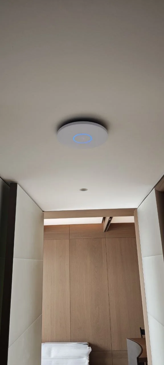 A ceiling with a round smoke detector or alarm and a partially open ceiling panel, viewed from a room with wood-paneled walls and a white towel or cloth folded on a surface.
