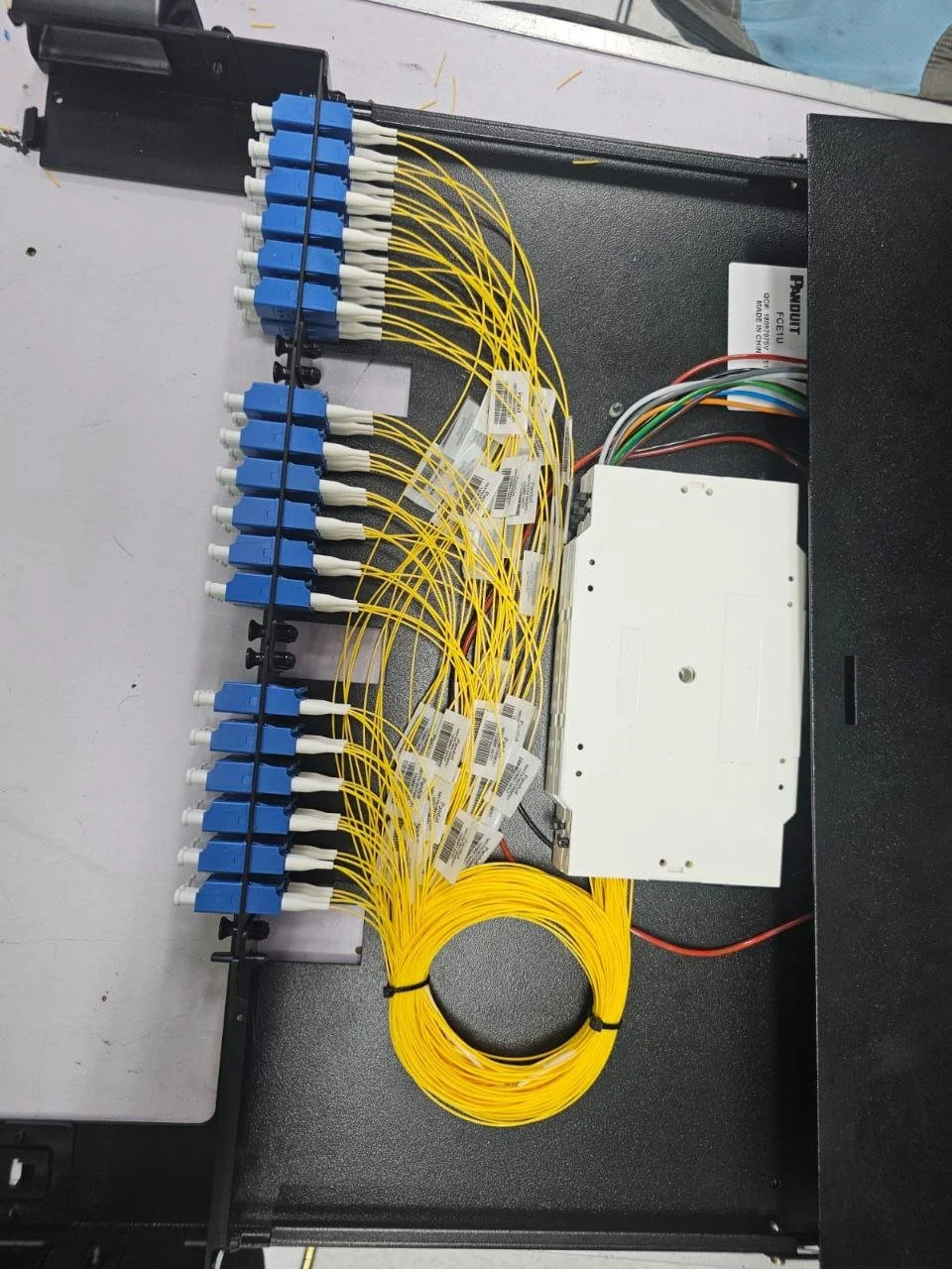 Fiber optic termination panel with yellow fiber optic cable, blue adapters, and a white electronic device, likely part of a telecommunications or data network setup.