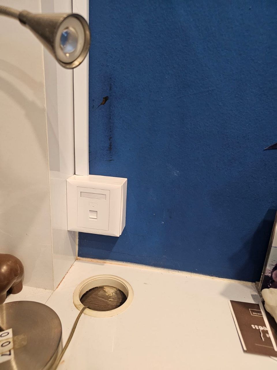 Close-up of a bathroom or kitchen counter with a metallic soap or lotion dispenser, an electrical outlet, a blue textured wall, and a hole in the countertop with a power cord inside.