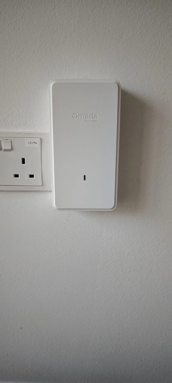 A white Omada by Etekcity Wi-Fi router mounted on a white wall next to a UK power socket.