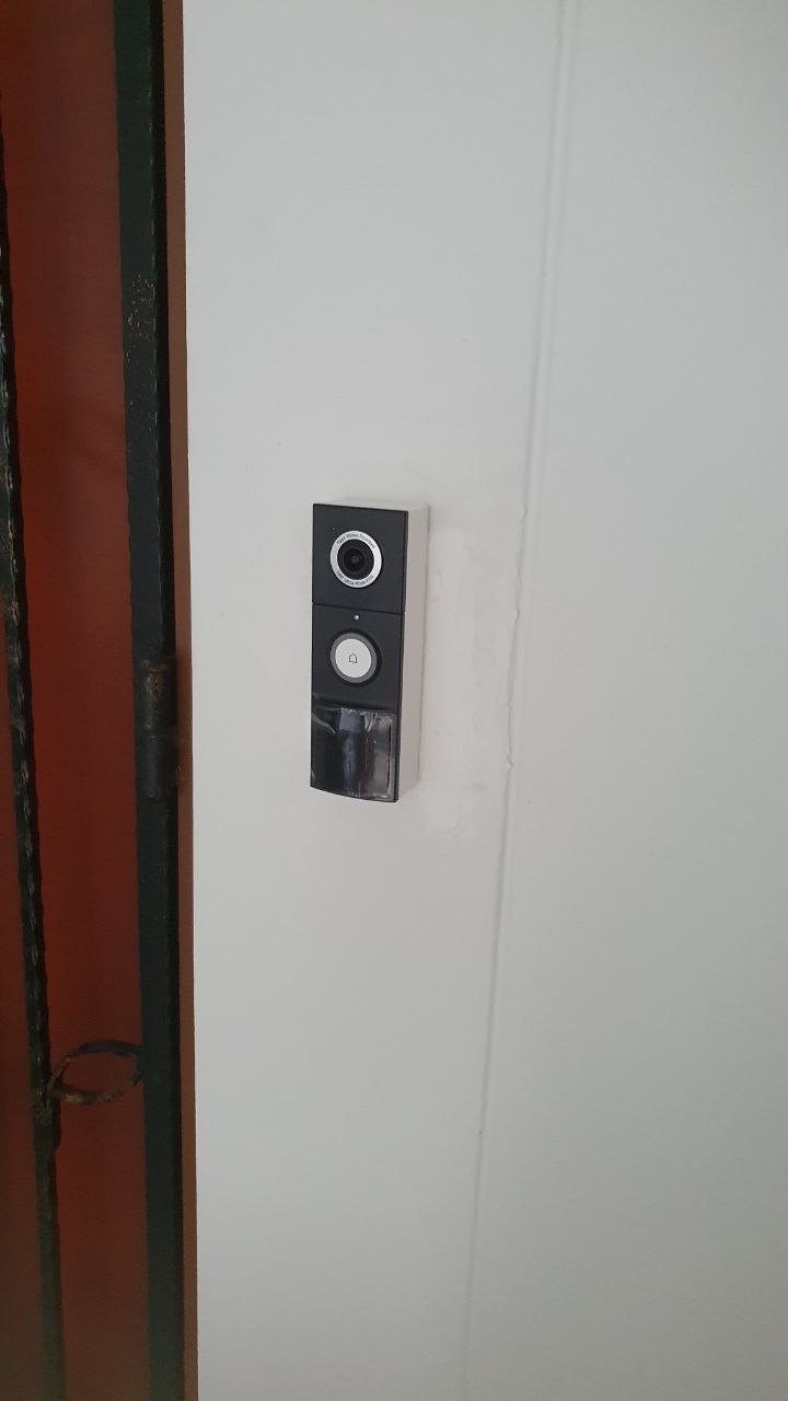 Doorbell camera with a camera lens at the top, a button in the middle, and a reflective surface at the bottom mounted on a white wall.