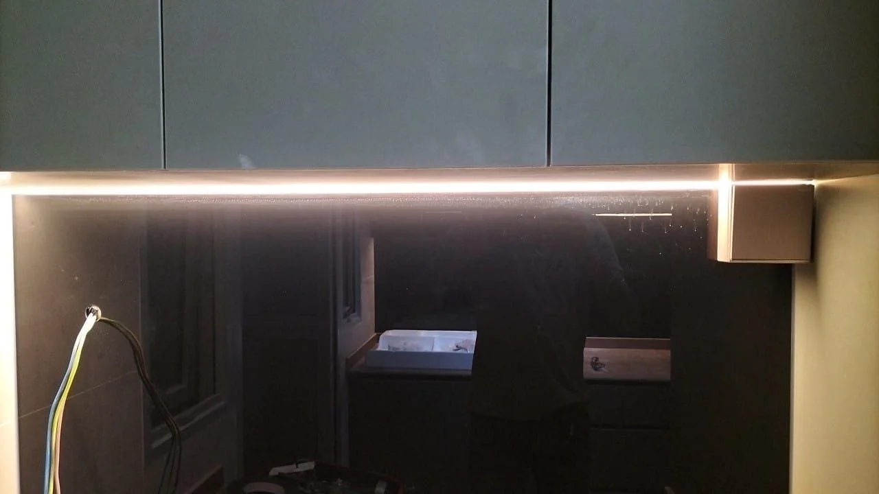 Close-up of a kitchen wall with cabinets, an LED light strip, and an incomplete electrical setup with wires hanging, and a mirror reflecting a person taking the photo.