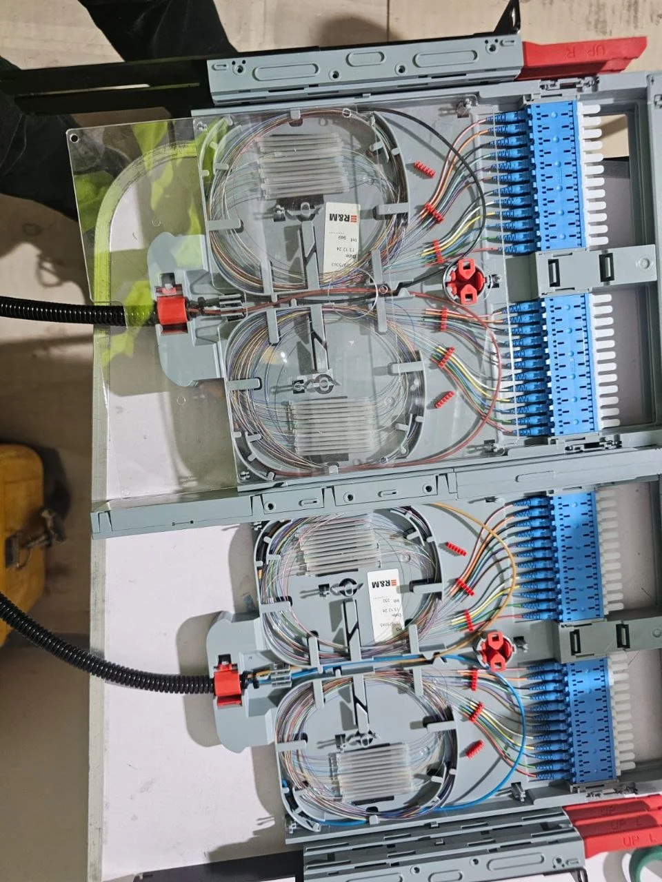 Inside an electrical fiber optic splice box, showing multiple fiber optic cables connected and organized with blue connectors and red spools, mounted on a white surface.