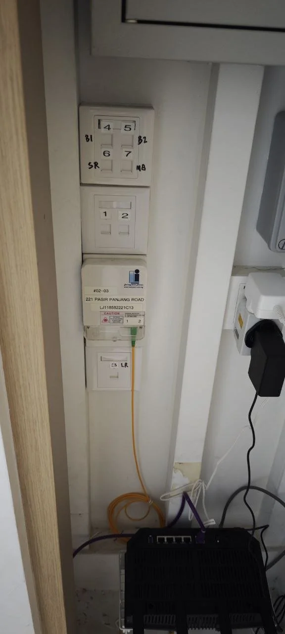 Electrical outlets, switches, and a power strip on a wall, with a black power strip or surge protector plugged into an outlet, and an orange Ethernet cable connected to a device on the floor.