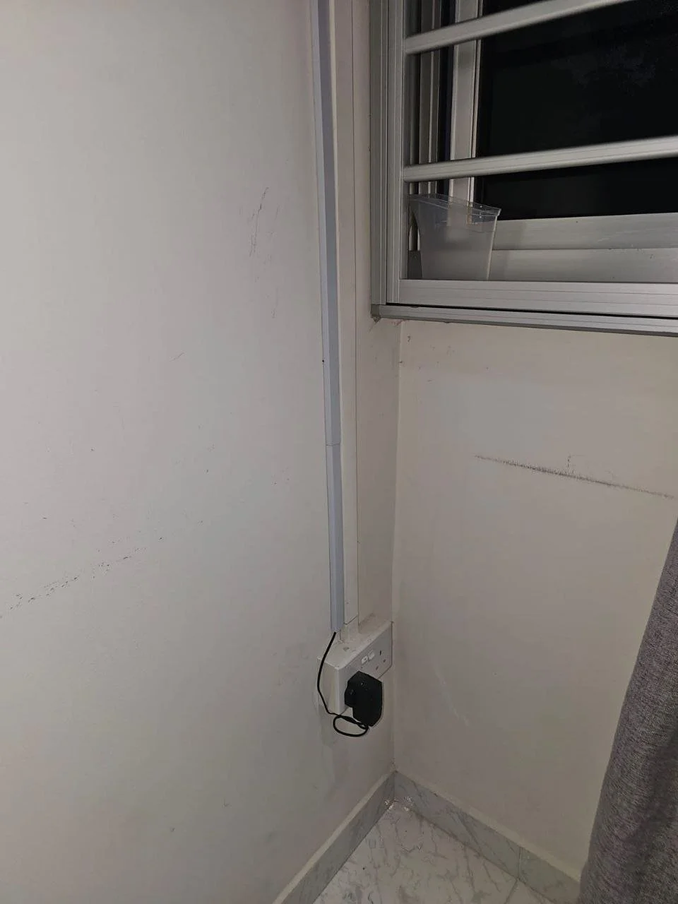 Corner of a room showing a power strip attached to the wall, with an electrical device plugged in. A window with a partially open metal shutter and a plastic container placed on the window sill.
