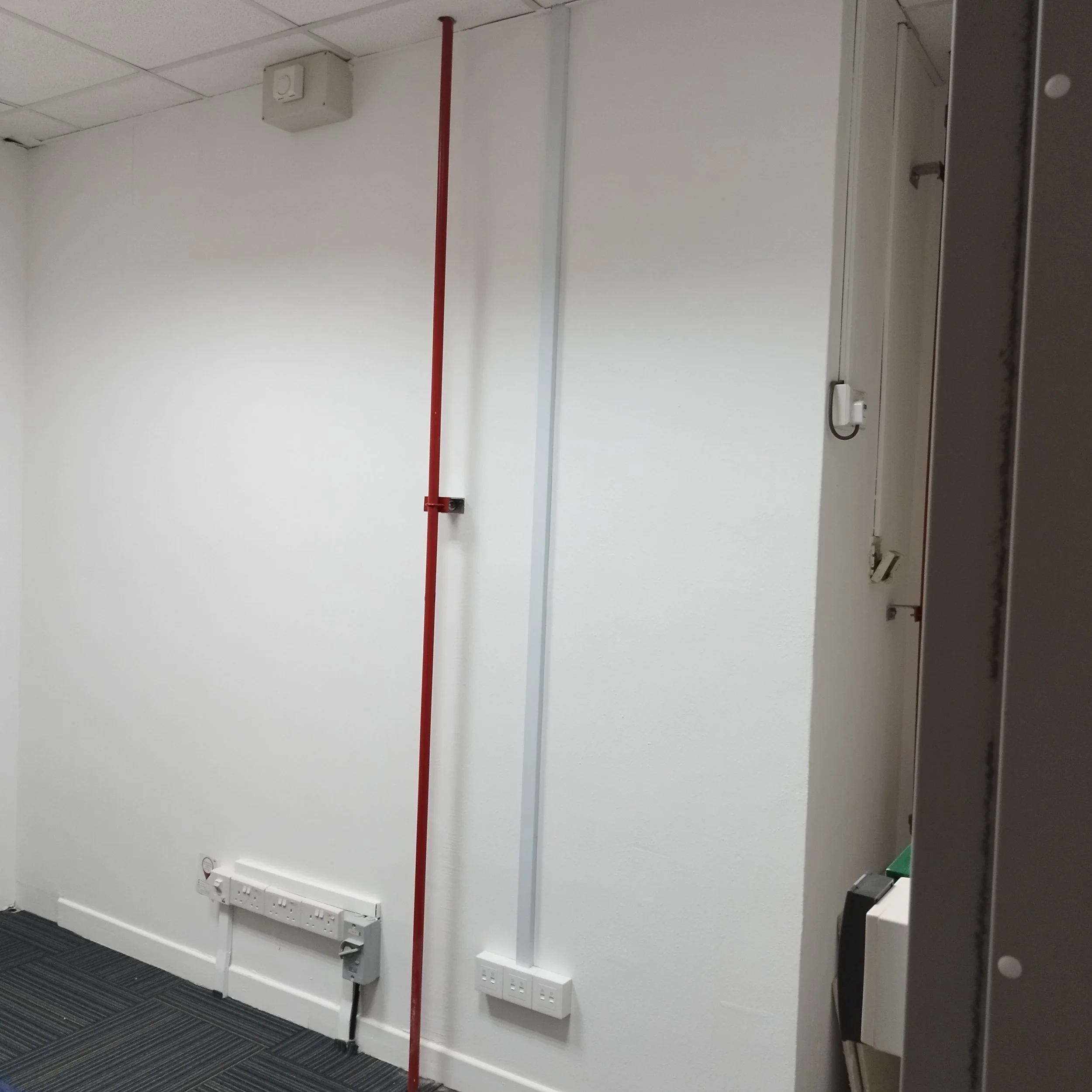 An empty office or utility room featuring a red emergency pull station on the wall, a white heater, and network sockets below it. The ceiling has suspended ceiling tiles, and there is a power outlet on the wall.