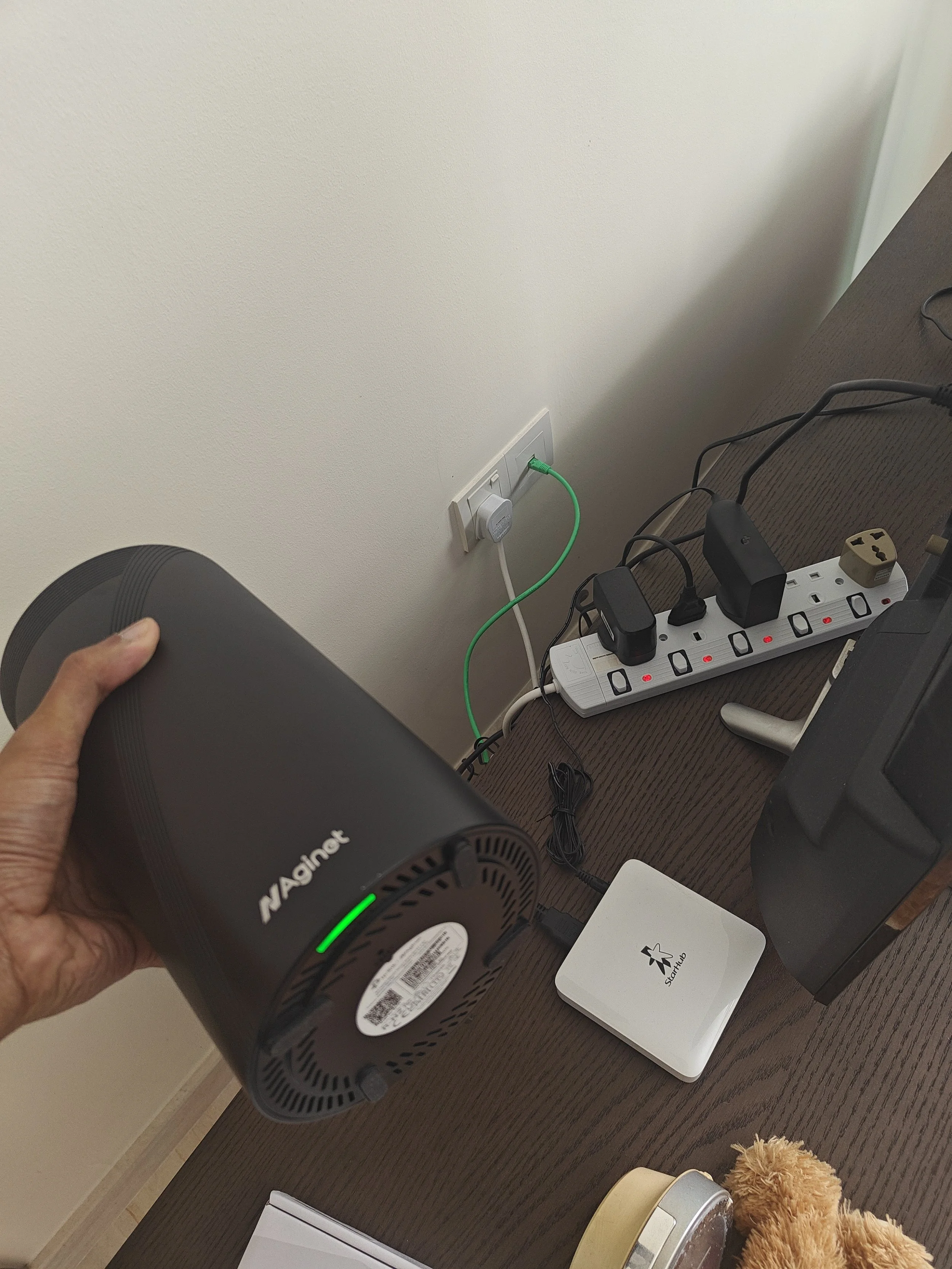 A person holding a black mini fan near a plug outlet on a beige wall. On the brown table below, there is a white power strip with plugged-in devices, a small white square device with a black star logo, a golden-colored container, a silver device, and