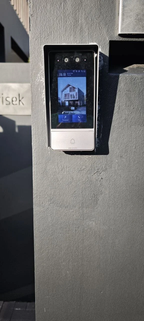 A digital intercom or access control device mounted on a gray wall, displaying a house image on its screen.