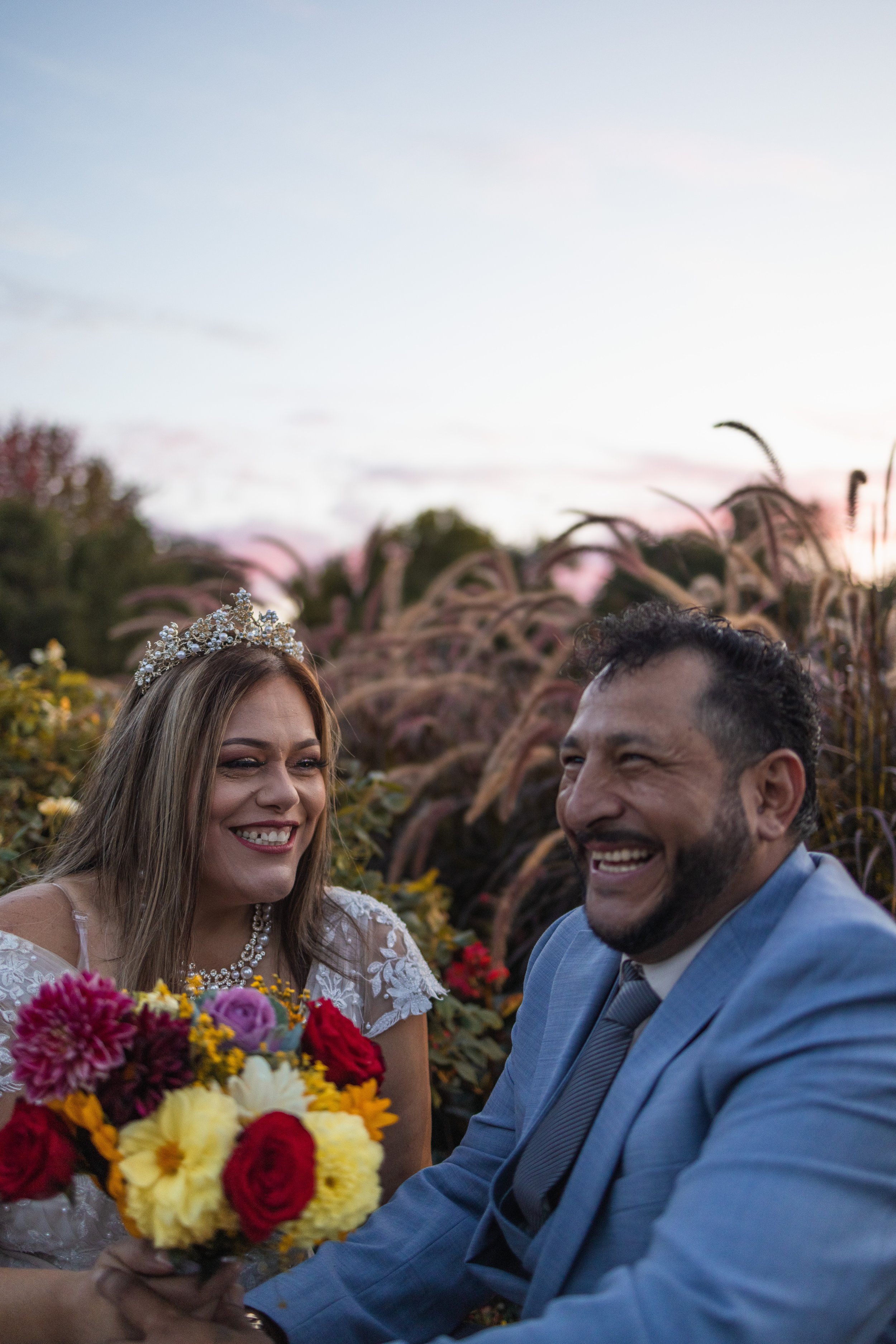 photograph of married couple laughing together in front of flowers and the sun setting