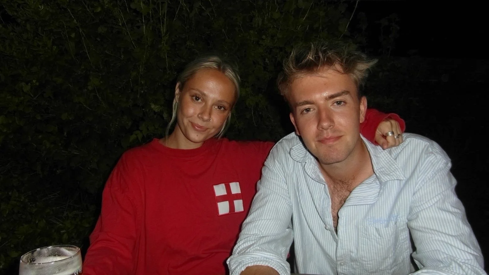Two young adults, a woman and a man, sitting outdoors at night. The woman has blonde hair, a nose piercing, and is wearing a red shirt with a white and red flag emblem. The man has light brown, tousled hair, and is wearing a white button-up shirt with the top buttons open. The woman has her arm around the man's shoulder, and they are both smiling slightly. There is a glass of beer on the table in front of them.