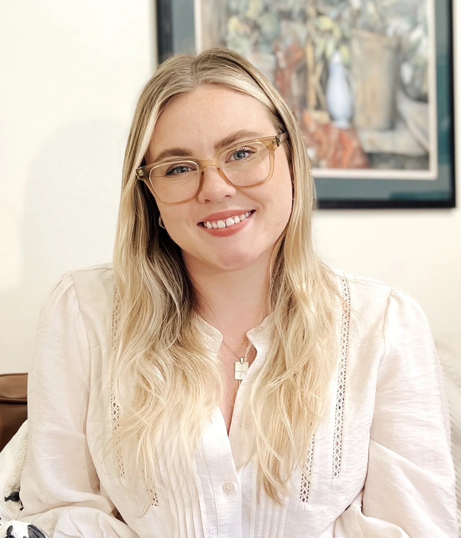 Color image of white woman with blonde hair and tan glasses smiling at camera with white blouse on