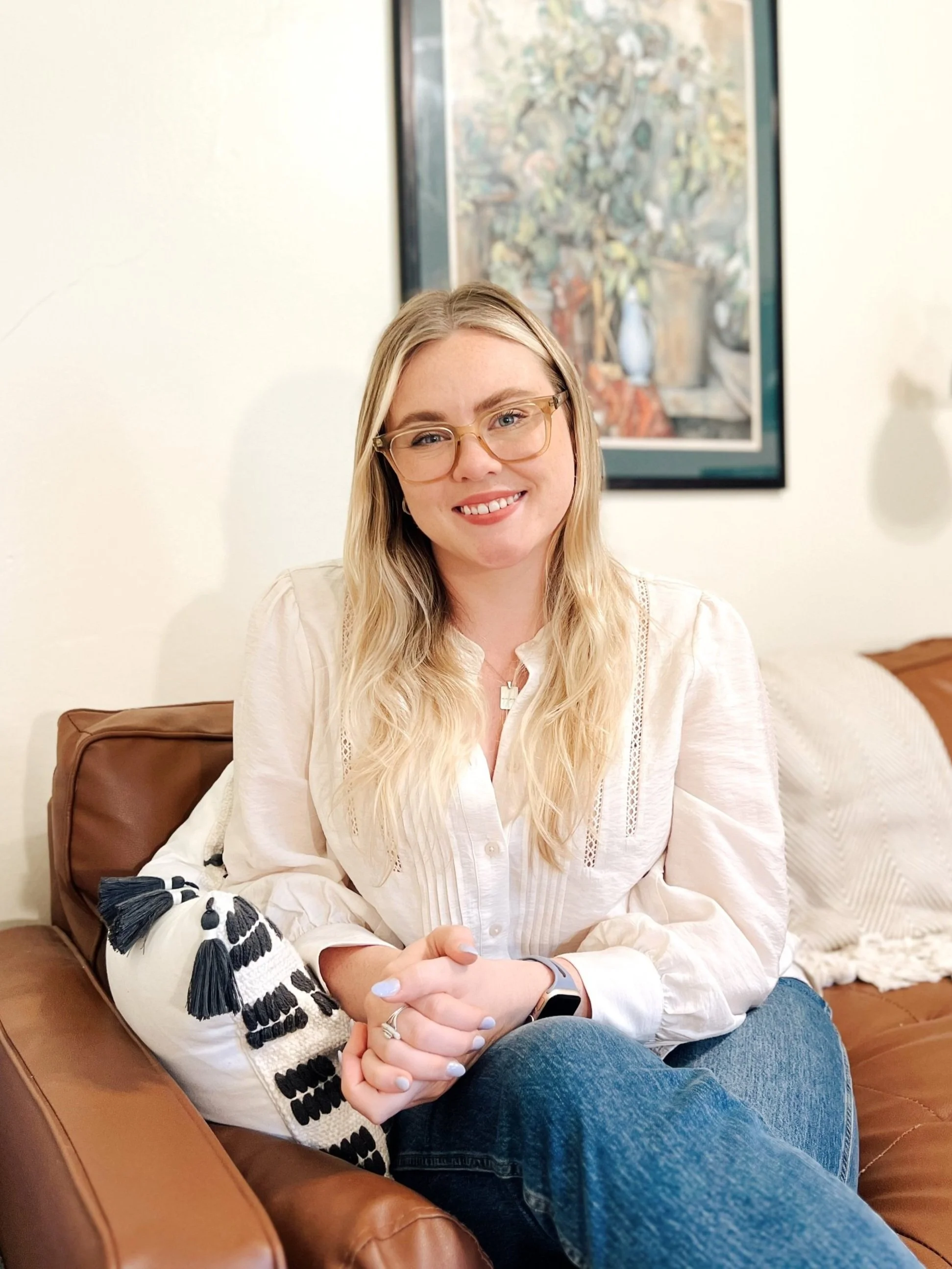 Image of Anika Vallejo (white, blonde, woman with glasses) wearing a white blouse and blue jeans on brown couch in front of a white wall