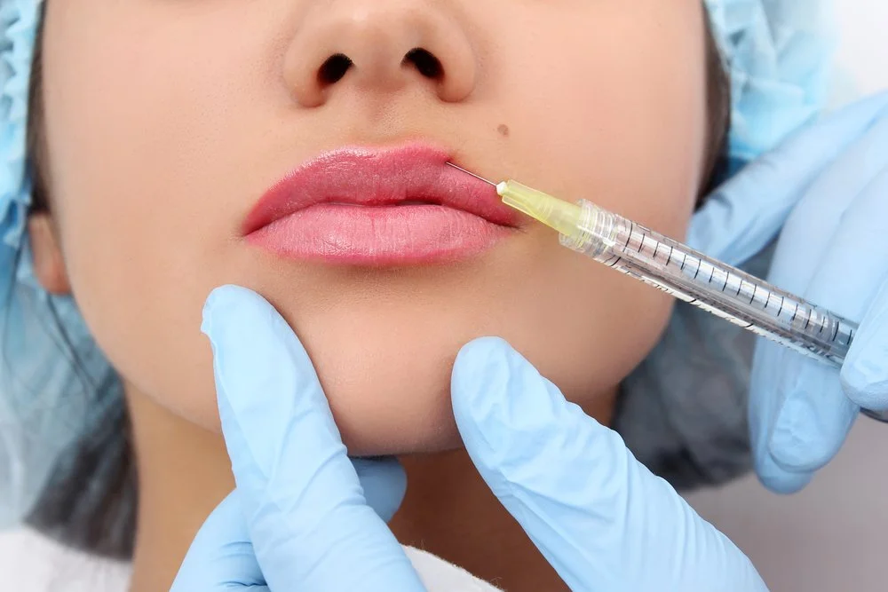 A person is receiving a cosmetic lip injection in a clinical setting, with gloved hands and a syringe near the lips.
